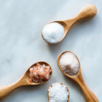 Various salt types in 4 wooden spoons on white marble.