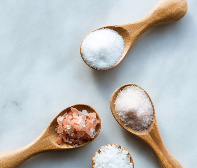 Various salt types in 4 wooden spoons on white marble.