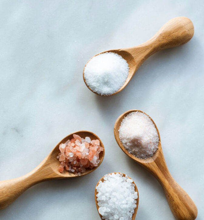 Various salt types in 4 wooden spoons on white marble.